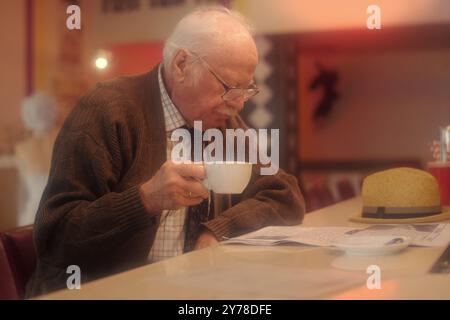 Elderly man with glasses, drinking coffee at cozy cafe and reading newspaper. Calm and peaceful scene with warm lighting evoking sense of relaxation and routine Stock Photo