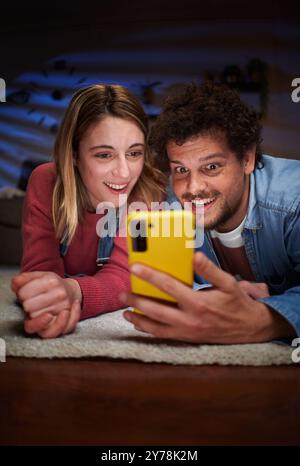 Vertical young Caucasian couple looking surprised at phone lying on carpet in living room at night. Stock Photo