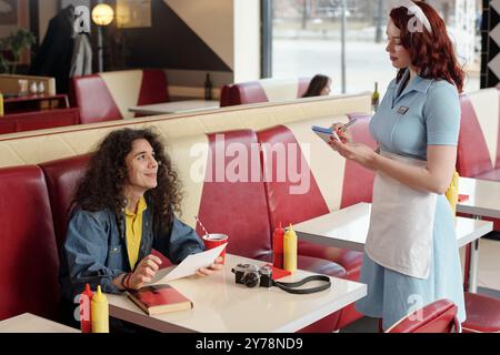 Smiling man with curly hair sitting at diner booth, interacting with waitress writing order on notepad, vintage camera and a notebook on table, ketchup and mustard dispensers Stock Photo