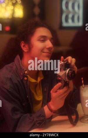 Young man sitting at table while holding vintage camera and enjoying moment drink in restaurant Inner setting features relaxed ambiance and smiling expression Stock Photo