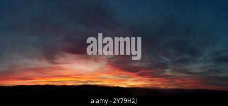 Stunning sunset paints sky with vibrant hues of red, orange, and pink, blending into deep blues and purples. Silhouetted hills create contrast against fiery horizon, adding drama to serene landscape. Stock Photo