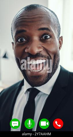 Surprised young African man in full suit looking at camera and smiling while having video call Stock Photo