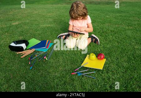 Kids study exam outside. Funny little student boy with tablet, Sit on lawn in park, studying at School backyard. Stock Photo