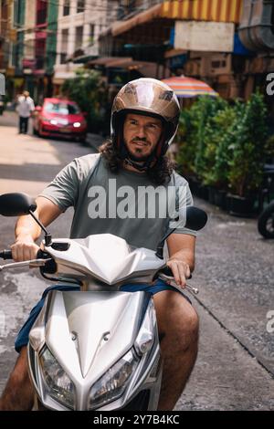 Vertical portrait of mid adult man driving a scooter with an open face helmet in the southeast of Asia; front view in a street of Sukhumvit, Bangkok, Stock Photo
