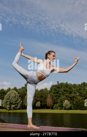 Professional yoga lover standing in dancer position and testing her body balance, she practicing yoga on river shore Stock Photo