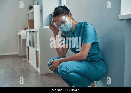 Tired young female nurse in protective workwear touching her head while working in the hospital Stock Photo
