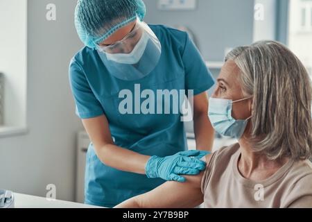 Female paramedic in protective workwear making coronavirus vaccination to mature woman while working in the hospital Stock Photo