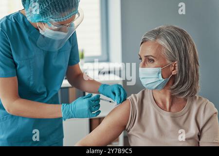 Female nurse in protective workwear making coronavirus vaccination to mature woman while working in the hospital Stock Photo