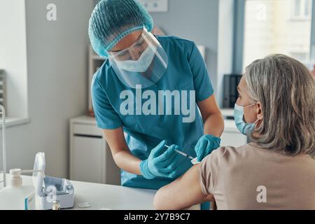 Female nurse in protective workwear making coronavirus vaccination to mature woman while working in the hospital Stock Photo