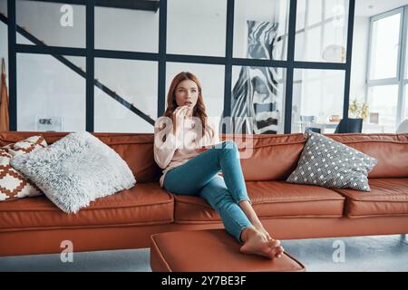 Surprised young woman watching television and eating popcorn while sitting on the sofa at home Stock Photo