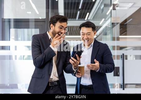Two colleagues sharing a light moment in office while watching a funny video on smartphone. Both men dressed in business suits, one holding a coffee cup, expressing laughter and surprise. Stock Photo