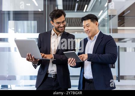 Two male professionals in business attire discussing project details using laptop and tablet. Conveying teamwork, communication, and technology in workplace. Ideal for concepts of collaboration Stock Photo