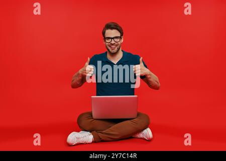 Happy young man in casual wear carrying computer and showing his thumbs up while sitting against red background Stock Photo