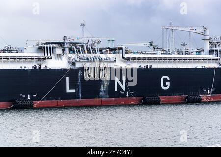 The lettering "LNG" is painted in large letters on the black hull of a bunker vessel at the jetty of the Mukran LNG terminal on the island of Rügen. White fenders and yellow mooring ropes hang at the ship's red waterline. Cranes and the piping scaffold of the regasification unit rise behind it into the cloudy sky. Spülfeld II, Sassnitz, Mecklenburg-Vorpommern, Germany. Stock Photo