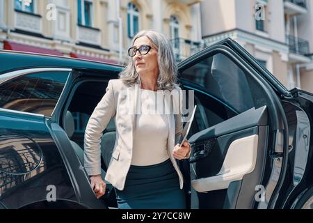 Mature beautiful woman in smart casual wear looking away while going out of the car outdoors Stock Photo