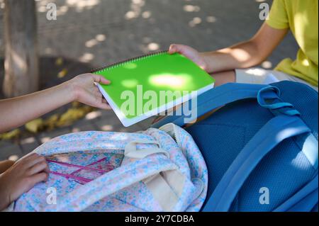 Two students share a green notebook outside, focusing on education and collaboration. Backpacks are visible. Stock Photo