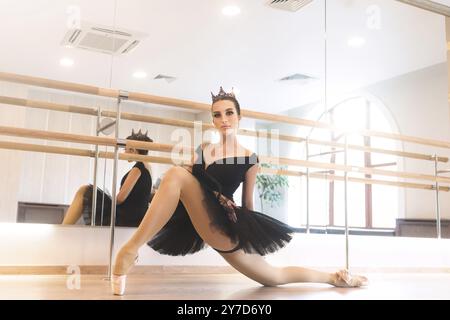 Ballet dancer in black tutu and pointe stretches on barre in ballet gym. elegant ballerina is standing near bar and mirror, preparing for perfomance Stock Photo