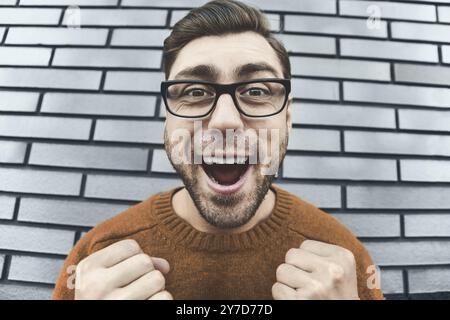 Young adult man over brown brick wall screaming proud and celebrating victory and success very excited, cheering emotion. Success concept Stock Photo