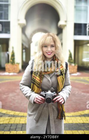 Blonde pretty photographer woman with retro camera in her hands while taking pictures at urban old architecture. Discover new places Stock Photo