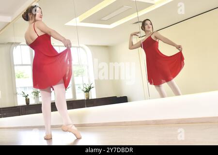 A young ballerina in a red dress and pointe shoes performs exercises in a dance studio Stock Photo