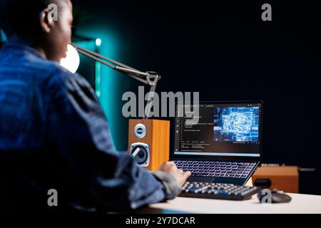 Professional female developer manages neural network at desk, working with code on laptop. Over the shoulder shot of african american freelancer developing cyber security, creating secure database. Stock Photo