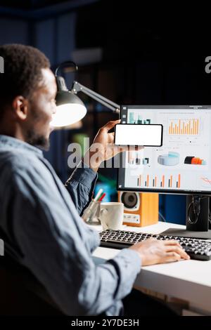 African american guy holds phone with white screen template at his workstation, using online analytics to solve business operations remotely on pc. Freelancer creating presentation, blank display. Stock Photo