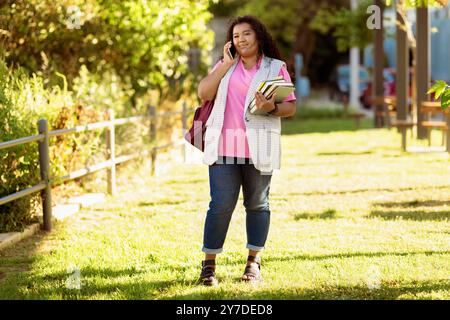Young woman in casual attire talking on a phone while holding books in a park Stock Photo