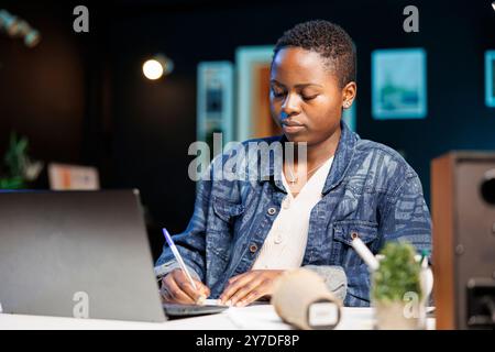 Focused black woman working from home, writing in notebook. African American freelancer using a laptop and multitasking, taking notes. Concentrated and determined to work efficiently. Stock Photo