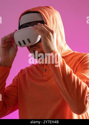 Man put on virtual reality headset in an orange hoodie against a vibrant pink backdrop engaging in immersive technology experience. Stock Photo