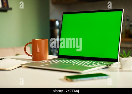 Modern home workspace featuring laptop with green screen, smartphone, coffee mug and small potted plants. Home office for freelancer with kitchen in b Stock Photo
