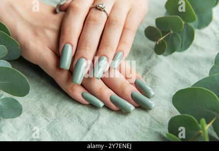 Beautifully manicured hands showcase long, green nails adorned with a delicate ring. The setting features soft eucalyptus leaves, creating a serene am Stock Photo