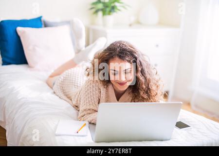 University student attending morning online class on laptop, sitting on bed in dorm room. Stock Photo