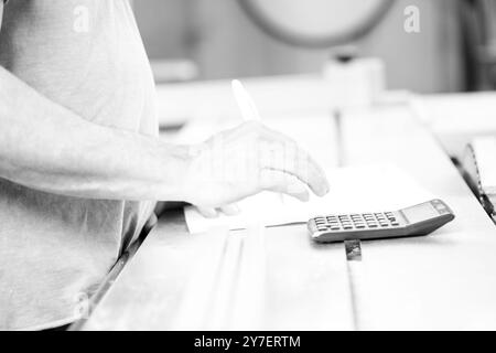 Carpenter is calculating costs in his workshop using a calculator and writing on a sheet of paper. - black n white - Stock Photo