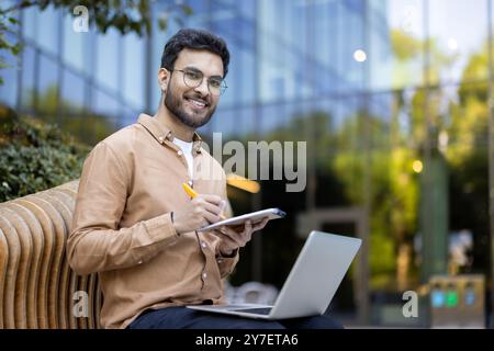 Young man smiling using laptop outdoors. Represents remote work, technology, productivity, and modern lifestyle. Ideal for depicting freelancer, entrepreneur, or student engaged in work or study. Stock Photo