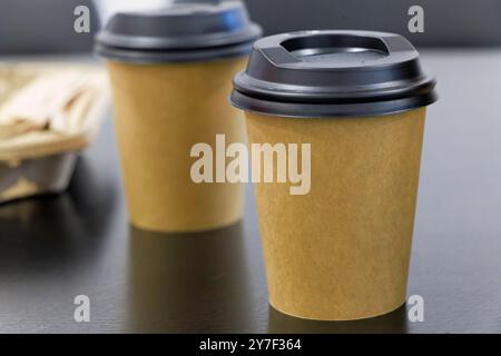 Two coffee cups with lids sit on a table. The cups are made of paper and have a black lid. Mockup Stock Photo
