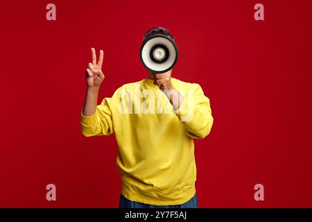 Person in bright yellow sweatshirt holds megaphone to face, making peace sign with their left hand against bright red background. Stock Photo