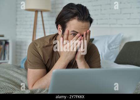 Terrified young man lying in bed and covering face with hand while looking at laptop Stock Photo