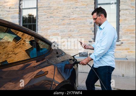 Man using smartphone while charging EV electric vehicle outside.. Stock Photo