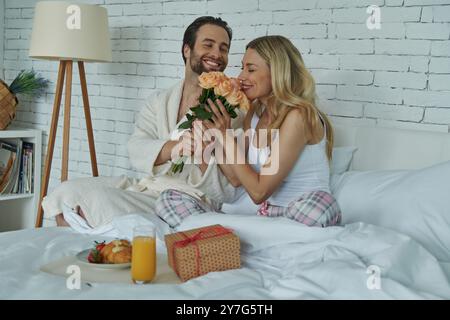 Cheerful man giving a bunch of roses to his girlfriend while sitting in bed together Stock Photo