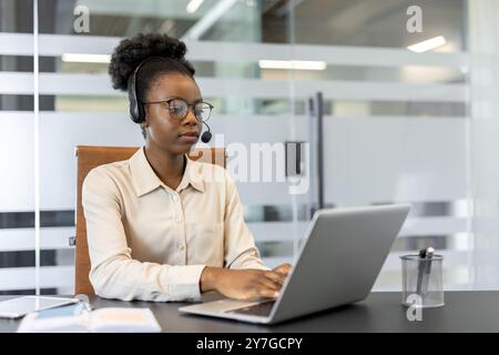 Confident woman wearing headset using laptop in modern office setting. Focused on customer service with phone support, displaying professionalism and dedication in workplace environment. Stock Photo