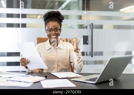 Smiling businesswoman sitting at office desk holding documents demonstrating success. Woman expressing confidence and achievement with clenched fist gesture. Stock Photo