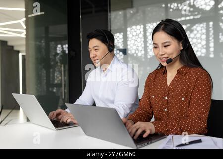 Two office professionals wearing headsets working on laptops in modern workspace. Collaboration, communication, and teamwork highlight their focus on customer service and telecommuting solutions. Stock Photo