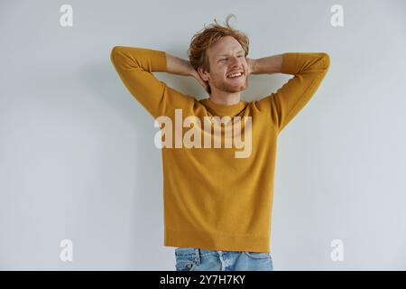 Relaxed young redhead man holding hands behind head while standing against wall Stock Photo