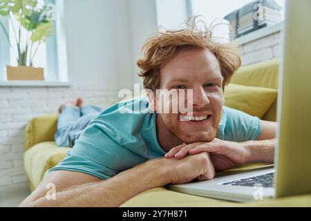 Cheerful redhead man using laptop and smiling while lying on the couch at home Stock Photo