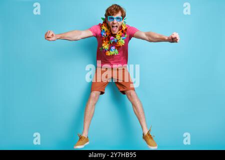 Cheerful redhead man in Hawaiian necklace jumping against blue background Stock Photo