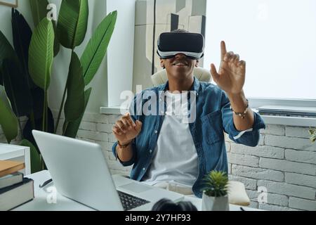 Joyful mixed race man wearing virtual reality glasses while sitting at working place Stock Photo