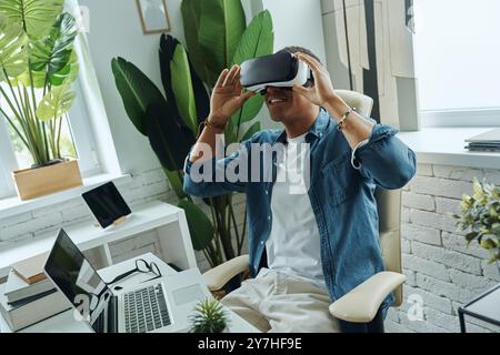 Cheerful mixed race man wearing virtual reality glasses while sitting at the desk Stock Photo