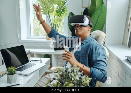 Joyful mixed race man wearing virtual reality glasses and gesturing while sitting at the desk Stock Photo
