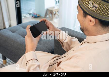 A young Muslim man with a smartphone in the living room at home Stock Photo