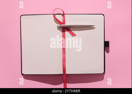 An open notebook with grid pages and a pen resting on top, set against a bright pink background featuring a red ribbon bookmark. Ideal for writing or Stock Photo